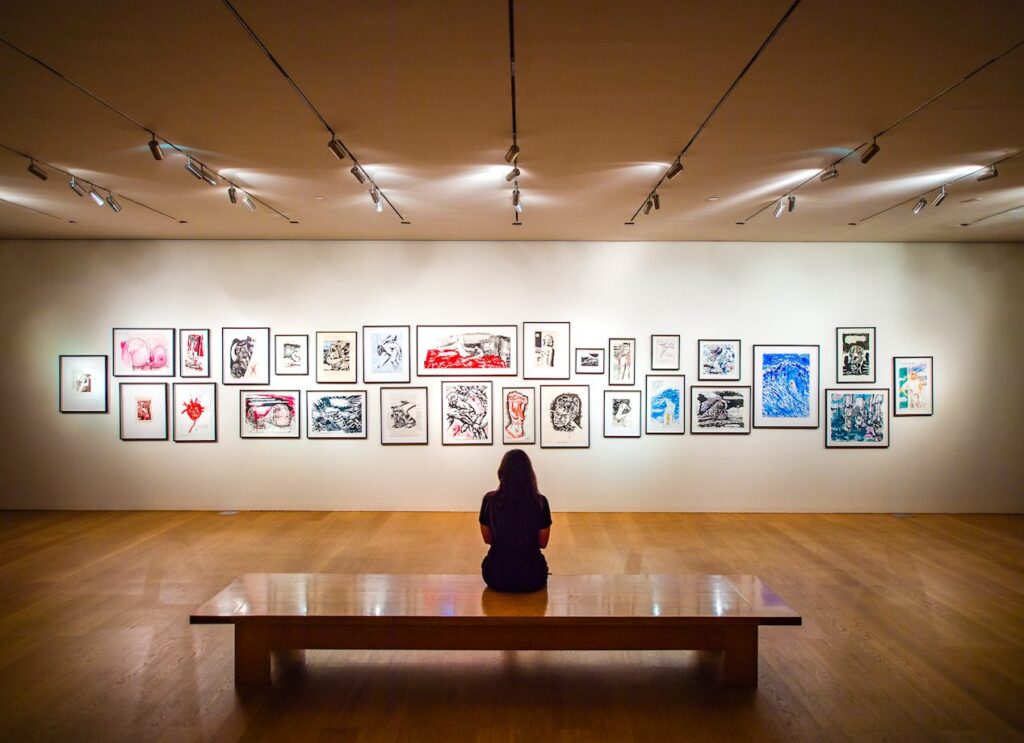 This photograph depicts a woman sitting on a bench and looking at a collection of paintings that are hanging on a wall in front of her. Perhaps she is contemplating if she wants to buy any of the art.
