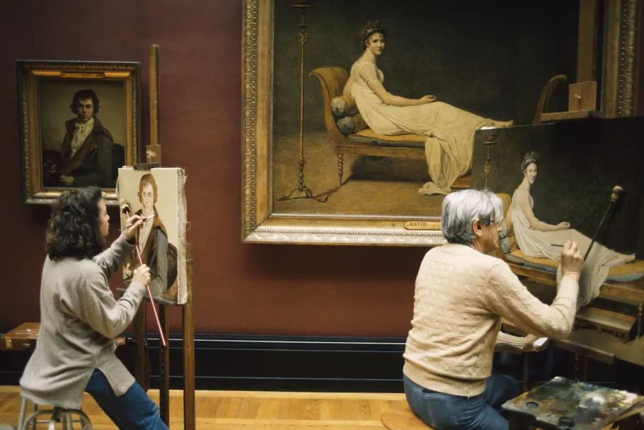 This photograph shows two people copying the paintings hanging on a wall in a museum.