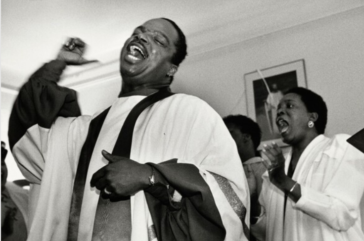 A Black man in gospel attire sings and motions to the air with his hands. Behind him, a Black woman in gospel attire sings along.