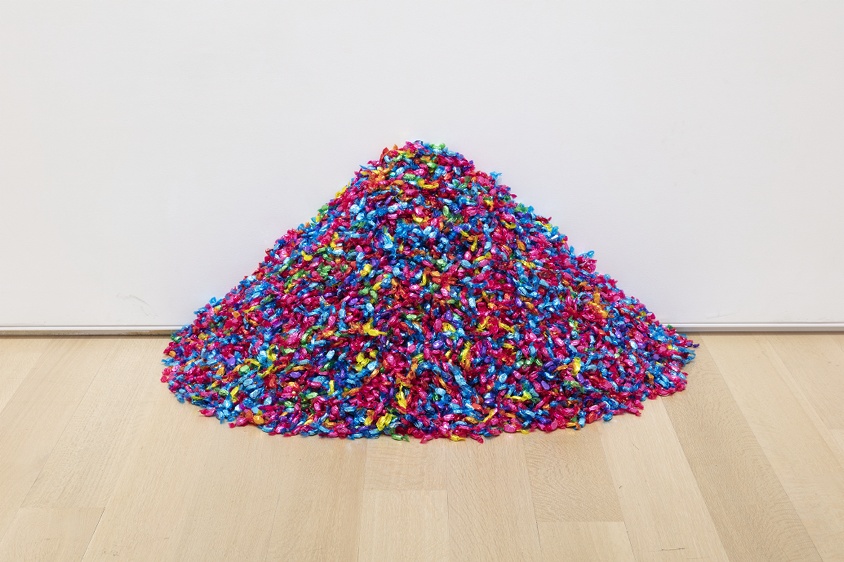 A pile of candies in multicolored wrappers sits on a wooden floor in front of a white wall.