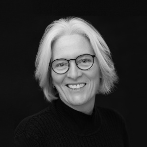 A black and white portrait of a white woman with white hair and black-rimmed glasses.