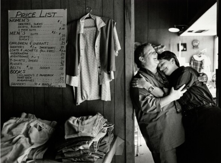 Two women embrace each other in a doorway. To their left is a sign listing prices for different articles of clothing.