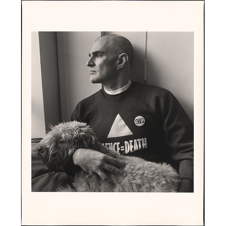 In a black and white photo, Larry Kramer, a bald white man, wears a sweater bearing a triangle and the words "Silence = Death". He holds a shaggy dog, Molly, and looks out a window to the left.