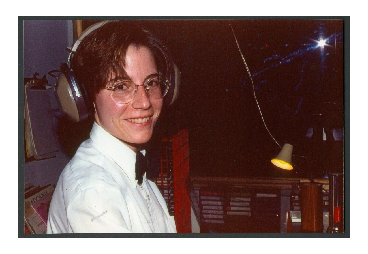 A white woman with short hair and a pair of over-the-ear headphones looks into the camera. She wears a white button-down shirt with a black bow tie. Behind her are crates of cassette tapes and vinyl records.