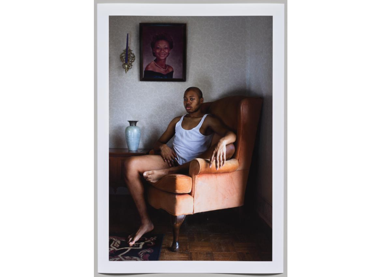 A Black person wearing only a white tank top sits in an armchair and looks into the distance. Their hand points toward their groin. On the wall above them is a portrait of a young woman.